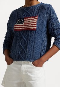 Blauer Strickpullover mit strukturiertem Zopfmuster und aufgestickter amerikanischer Flagge auf der Vorderseite. Grob gestricktes Design mit lockerem Sitz.