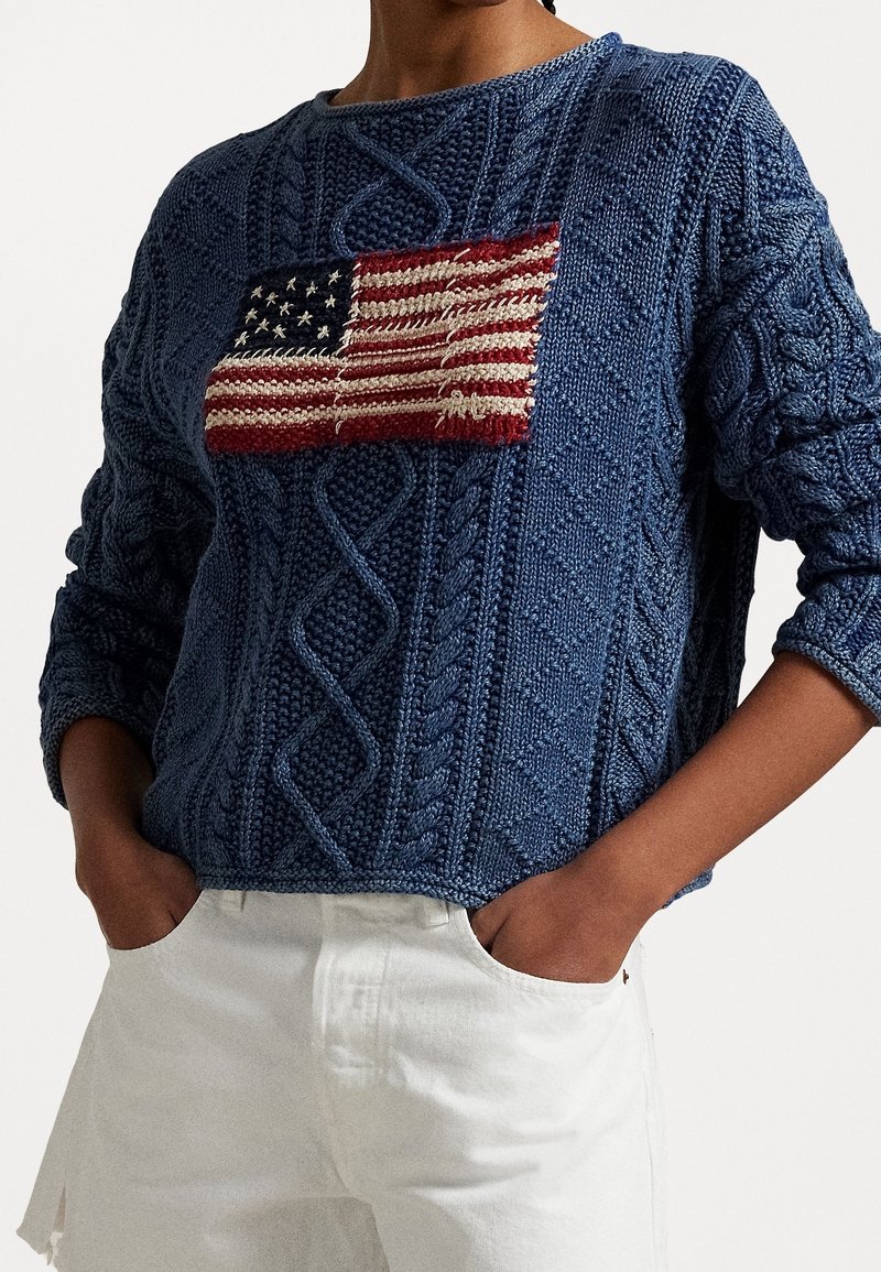 Blauer Strickpullover mit strukturiertem Zopfmuster und aufgestickter amerikanischer Flagge auf der Vorderseite. Grob gestricktes Design mit lockerem Sitz.