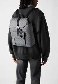 Sac à dos gris à motifs avec des bretelles noires, une fermeture magnétique et une poche extérieure. Porté sur un pull sombre et un pantalon gris.