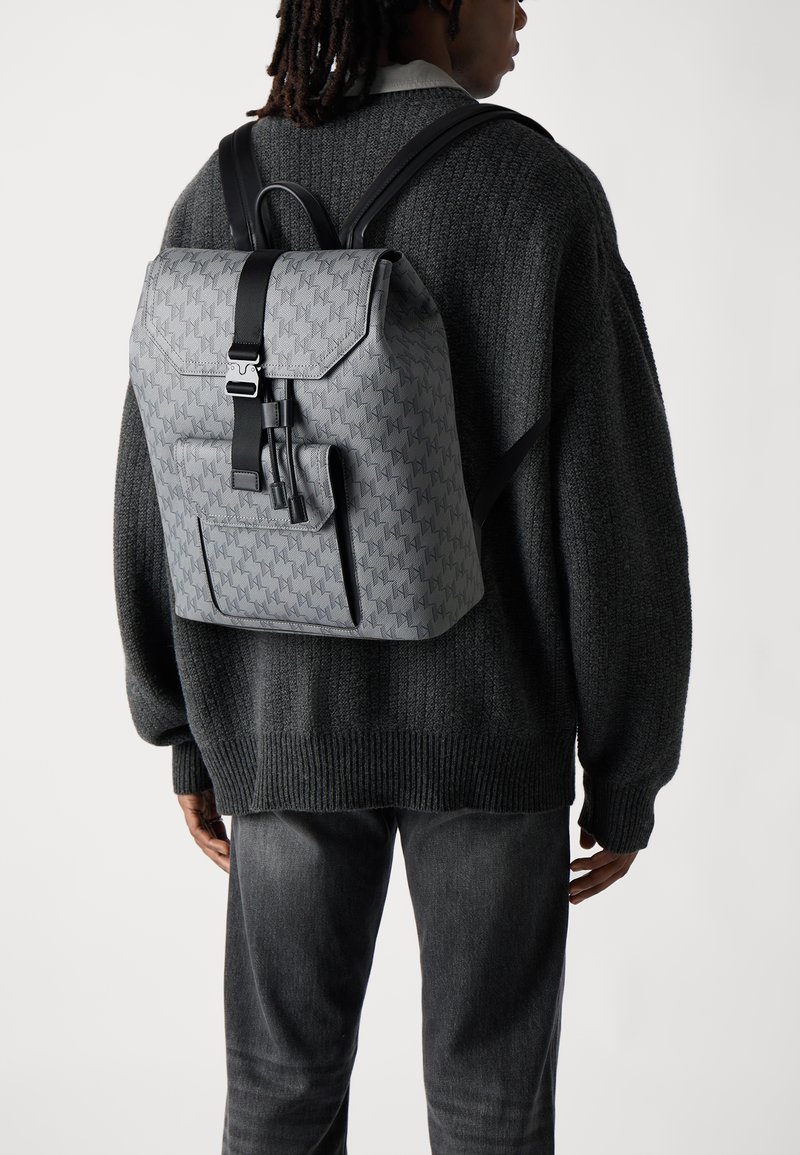 Sac à dos gris à motifs avec des bretelles noires, une fermeture magnétique et une poche extérieure. Porté sur un pull sombre et un pantalon gris.