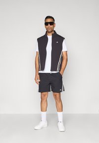 Chaleco sin mangas negro con rayas blancas, camiseta blanca y pantalones cortos negros con rayas laterales, combinados con zapatillas blancas y gafas de sol.