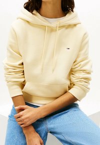 Vaaleankeltainen huppari, jossa on kenguru-tasku ja nyörihuppu. Ribbaatut hihansuut ja helma, jossa on pieni logo rinnassa. Pehmeä kankaan texture.