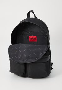 Sac à dos en nylon noir avec deux poches avant zippées et une doublure intérieure ornée du logo "HUGO" en répétition. La rabat ouvert révèle un intérieur spacieux.