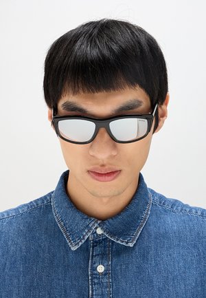 Giovane uomo con capelli corti e neri indossa occhiali da sole specchiati con montatura nera e una camicia di denim blu con bottoni su sfondo uniforme.