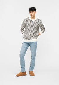 Jeune homme debout, les mains derrière le dos, portant un pull crème à motifs, un jean bleu clair et des bottines marron, sur fond blanc.