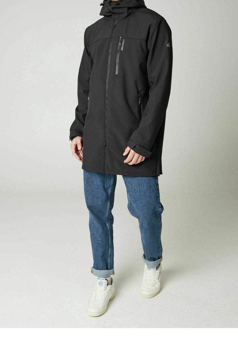 Homme portant une veste noire à capuche avec des poches à fermeture éclair, un jean bleu retroussé et des baskets blanches, debout devant un fond uni.