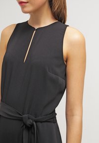 Svart ärmlös jumpsuit med hög halsringning, som har en liten nyckelhålskärning och en knutdetalj i midjan för en strukturerad silhuett.