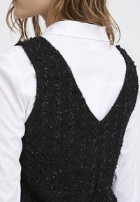 Crna haljina s teksturom i dubokim V-dekolteom na leđima, ukrašena valovitim uzorcima i suptilnim šljokicama; nosi se preko bijele košulje s ovratnikom.