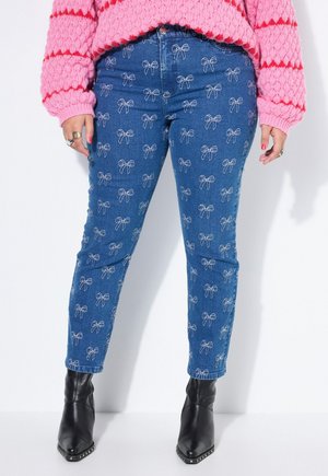 Person, der bærer blå jeans med hvide sløjfe-mønstre, en pink striktrøje med røde striber, sorte spidse støvler og ringe på fingrene.