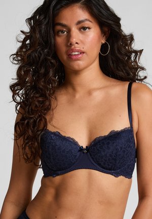 Femme avec des cheveux noirs bouclés portant un soutien-gorge en dentelle bleu marine et de grandes boucles d'oreilles en anneaux, sur un fond clair uni.