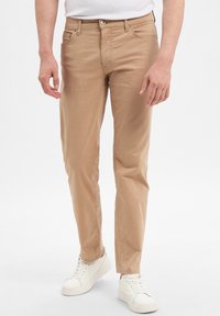 Pantaloni in cotone beige a taglio slim, con zip, chiusura a bottone e tasche, abbinati a sneakers bianche su uno sfondo neutro.