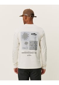 Pitkähihaisessa kermaisessa paidassa on graafinen kuvio, joka sisältää abstrakteja muotoja ja tekstiä, painettuna selkäpuolelle. Ruskea lippis täydentää asua.