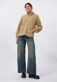 Jeune femme portant un pull en polaire beige, un jean large délavé et des bottes noires chunky, se tenant debout et souriant devant un fond uni.