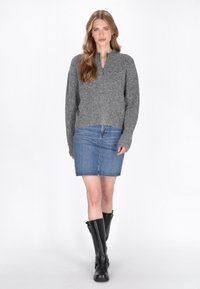 Pulover gri tricotat cu guler înalt, asortat cu o fustă din denim. Modelul poartă cizme negre înalte. Design simplu, curat, pe un fundal alb.