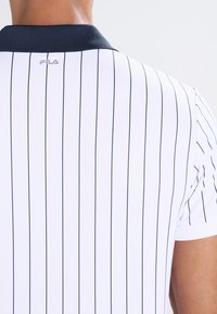 Hvit polo skjorte med marineblå vertikale striper, ribbet krage og en liten logo på baksiden. Laget av pustende, strukturert stoff.