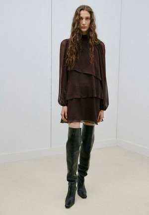 Mujer con cabello largo y ondulado, vestida con un vestido de capas marrón oscuro y botas negras hasta la rodilla, de pie frente a paredes lisas de color claro.