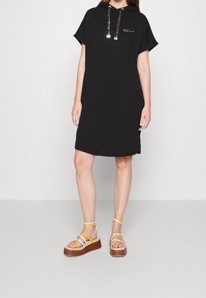 Femme portant une robe à capuche noire mi-longue aux manches courtes et des sandales compensées avec des brides blanches et jaunes.