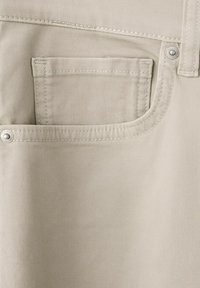 Beige denim fabric close-up showing front pocket with coin pocket, topstitching, rivets, and belt loop detail.