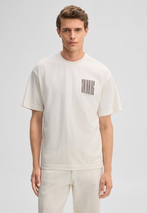 Junger Mann mit kurzen lockigen Haaren, der ein hellbeiges T-Shirt mit dem Text "SWIMMING" auf der Brust trägt und vor einem einfarbigen grauen Hintergrund steht.