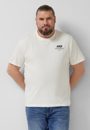 Mann mit Bart und zurückgebundenen Haaren, der ein weißes T-Shirt mit dem Aufdruck "ARR Rethink&Reload" und blaue Jeans trägt, steht vor einer grauen Wand.