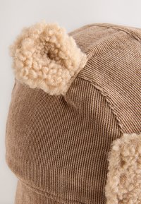 Rjava klobuk iz korduroja z mehkim bež poudarkom na ušesih. Ima teksturirano površino in zaokroženo obliko z vidnimi šivi.