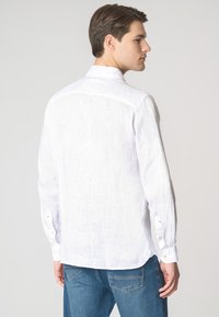 Camicia bianca di lino a maniche lunghe con colletto, dotata di chiusura con bottoni e orlo leggermente curvo, indossata con jeans blu.