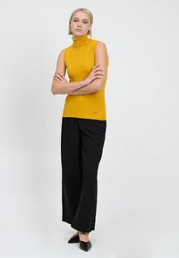 Geel mouwloos coltruitje met ribbelstructuur, gecombineerd met zwarte wijde broek en scherpe zwarte schoenen.