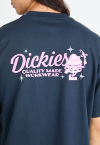 Tamsiai mėlynas medvilninis marškinėliai su dideliu rožiniu piešiniu, kuriame užrašas "Dickies" ir animacinis personažas, laikantis įrankį, dekoruotas žvaigždutėmis.