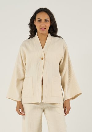 Vrouw die een beige textuurige jas draagt met brede mouwen, een enkele houten knoop, voorzakken en bijpassende broek, staand tegen een lichte achtergrond.