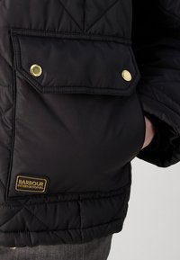 Casaco acolchoado preto com um bolso de botão de pressão dourado; apresenta um acabamento texturizado e um emblema da Barbour International na bainha.