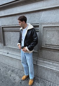 Νέος άνδρας με μαύρο μπουφάν από shearling, ανοιχτά τζιν και καφέ μπότες κρατώντας ένα φλιτζάνι καφέ, στέκεται μπροστά σε έναν αναλυτικό πέτρινο τοίχο.