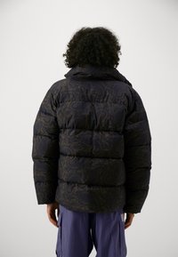 Giacca a vento trapuntata blu navy con motivo paisley, cappuccio e polsini elastici, abbinata a pantaloni cargo viola. Vista posteriore mostrata.