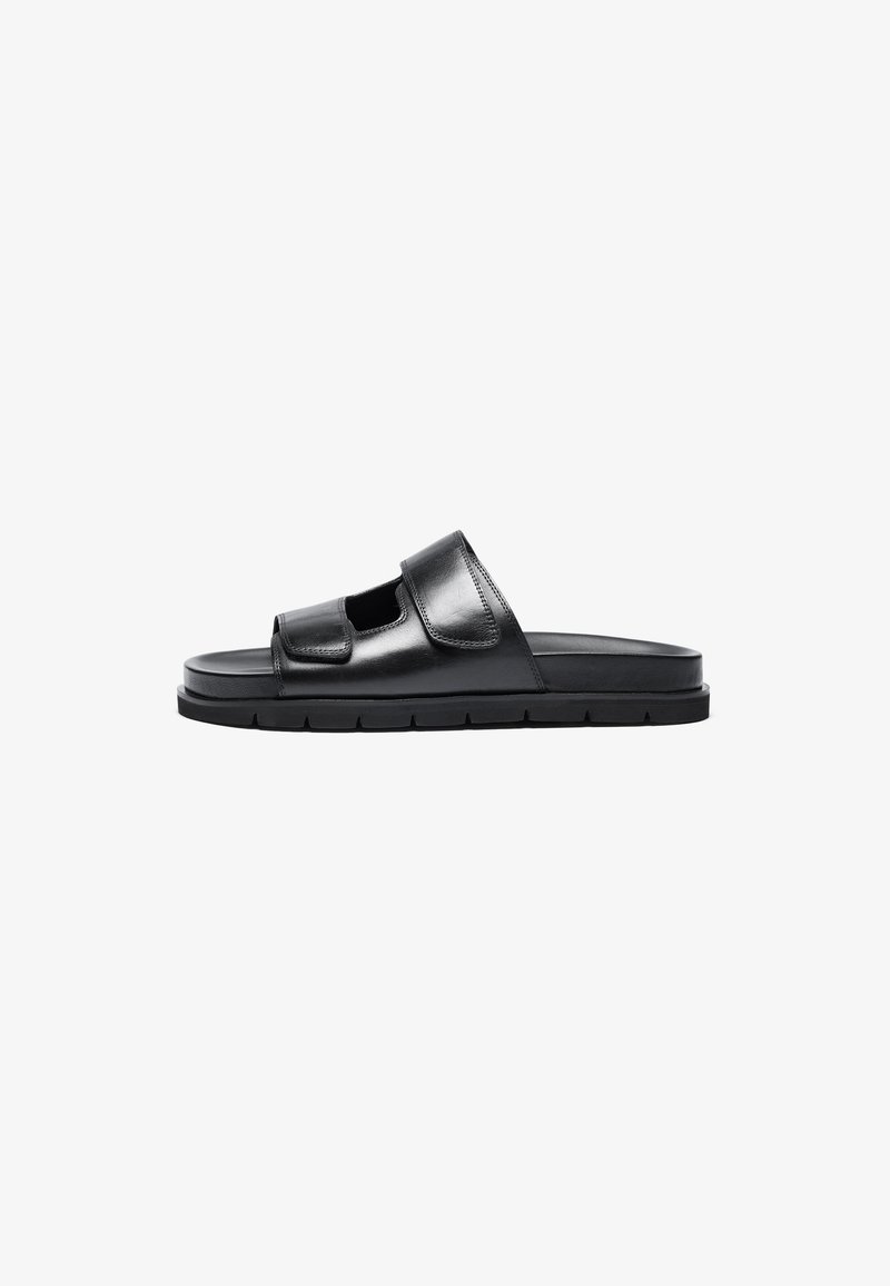 Black leather slide sandal with a wide adjustable strap, open toe, and textured sole. Minimalist design with a flat, cushioned footbed.