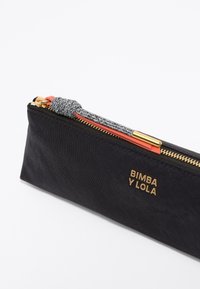 Bolsita negra de tela. Tiene un zipper dorado, un cordón gris decorativo y el logo "BIMBA Y LOLA" en dorado. Forma rectangular. Diseño sencillo.