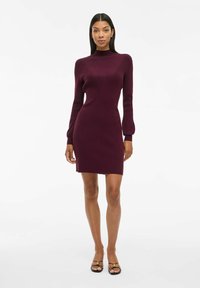 Robe tricotée bourgogne avec un col haut et de longues manches bouffantes. Présente une silhouette cintrée et une légère fente à l'ourlet pour faciliter le mouvement.