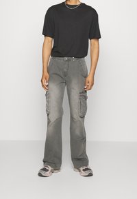 Graue Cargo-Hosen mit lockerem Schnitt, ausgestattet mit mehreren Taschen, einer ausgewaschenen Textur und dezenten Nahtdetails. Kombiniert mit einem schwarzen T-Shirt.