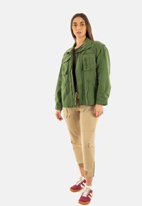 Chaqueta utilitaria verde con cuatro bolsillos frontales, combinada con pantalones cargo beige que tienen múltiples bolsillos, y zapatillas rojas y blancas.