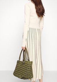 Bolsa de tote tejida en verde oliva con asas dobles, superficie texturizada y un diseño espacioso, combinada con una blusa crema y una falda midi a rayas.