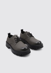 Zapatos con suela adherente en material sintético de color gris oscuro con acabado suave, punta redondeada y cordones negros. Incluye una tira en el talón.
