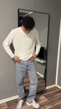 Νεαρό άτομο προσαρμόζει ανοιχτό μπλε τζιν, φορά άσπρο μακρυμάνικο πουκάμισο και αθλητικά παπούτσια, στέκεται μπροστά σε ένας καθρέφτη ολόσωμο μέσα σε ένα δωμάτιο.