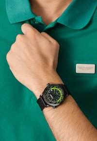 Reloj negro con una correa de goma texturizada, acentos verdes y una esfera oscura que muestra múltiples zonas horarias, llevado en una mano contra una camiseta polo verde.