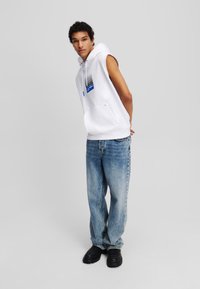 Felpa senza maniche bianca con design grafico blu; abbinata a jeans larghi di un blu chiaro e scarpe nere. Stile semplice e minimalista.