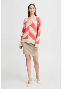 Pull beige à col en V avec un grand motif en chevrons rouge, associé à une jupe beige à côtes ajustée. Le mannequin porte des talons blancs pointus.