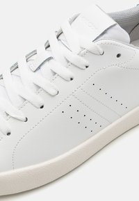 Hvite lær sneakers med en jevn tekstur, med perforeringer på siden, flate hvite lisser og en teksturert gummisåle.