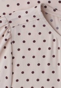 Tissu beige clair avec des pois brun foncé espacés régulièrement, présentant une couture froncée à l'épaule et une couture lisse à l'encolure.