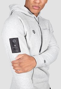 Light gray hoodie featuring a drawstring, black accents, and a branded patch on the left sleeve. Textured fabric with ribbed cuffs.