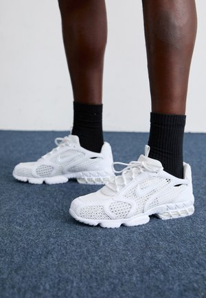 White athletic shoes with mesh upper, textured accents, and cushioned sole. Paired with black ribbed ankle socks on a blue carpeted surface.