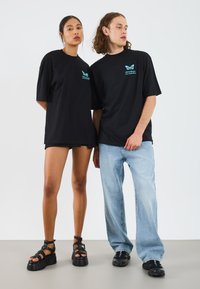 Sorte oversized t-shirts med turkist sommerfugl-logo, parret med lyseblå lige ben jeans. Fodtøjet inkluderer sorte platform sandaler.
