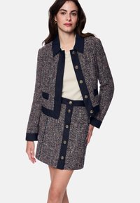 Tweed jacket and skirt set in navy, red, and cream. Features a collar, gold buttons, and side pockets on the jacket. Structured silhouette.