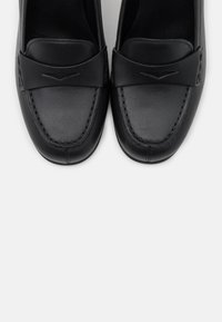 Mocassins à enfiler en cuir noir avec un détail de couture et une découpe en forme de V sur la bride supérieure, vus de dessus.